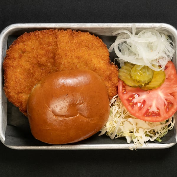 A crispy fried chicken sandwich with pickles, tomato, onions, and shredded lettuce on a metal tray.