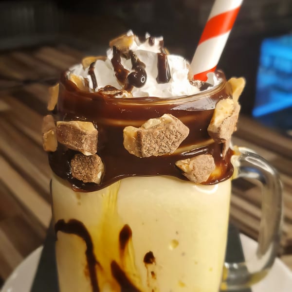 A Roadhouse milkshake in a mug, topped with whipped cream, chocolate drizzle, candy pieces, and a red-striped straw.