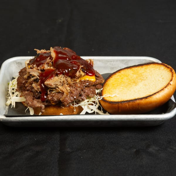 A Roadhouse burger with pulled pork, BBQ sauce, cheese, and shredded cabbage on a metal tray.